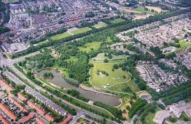 Schelfhorstpark,, Luchtfoto van groot stedelijk park omgeven door woonwijken, met grasvelden, bosrijke secties en een groot vijver met kronkelende kustlijn. Paden en gebouwen zichtbaar in het park, agenda World of Veterans